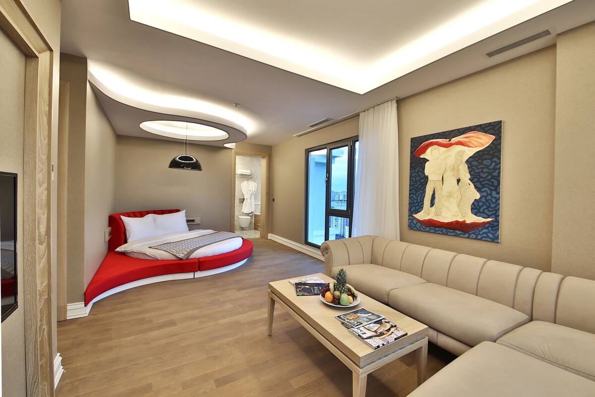 A modern hotel room with a curved bed, a sofa, a coffee table with magazines, and a large window with a view.