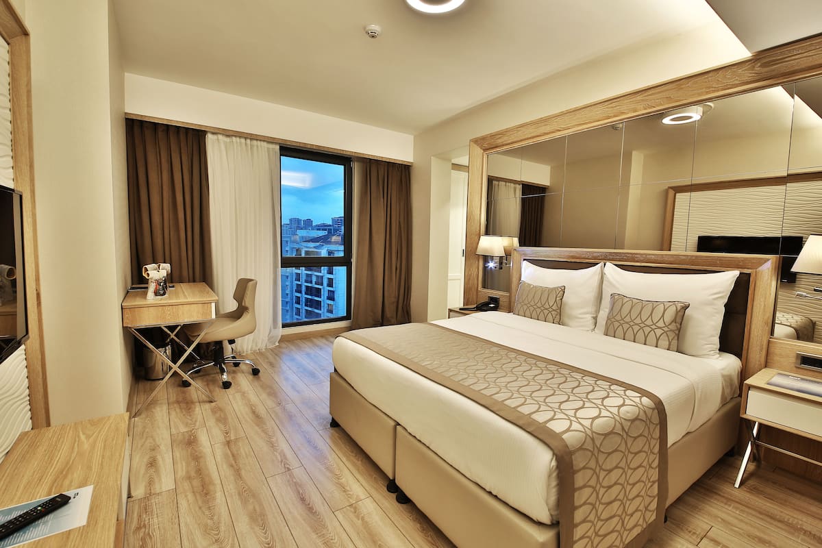 A modern hotel room with a large bed, a desk, and a chair. There is a large window with curtains, a mirror, and a view of the city.