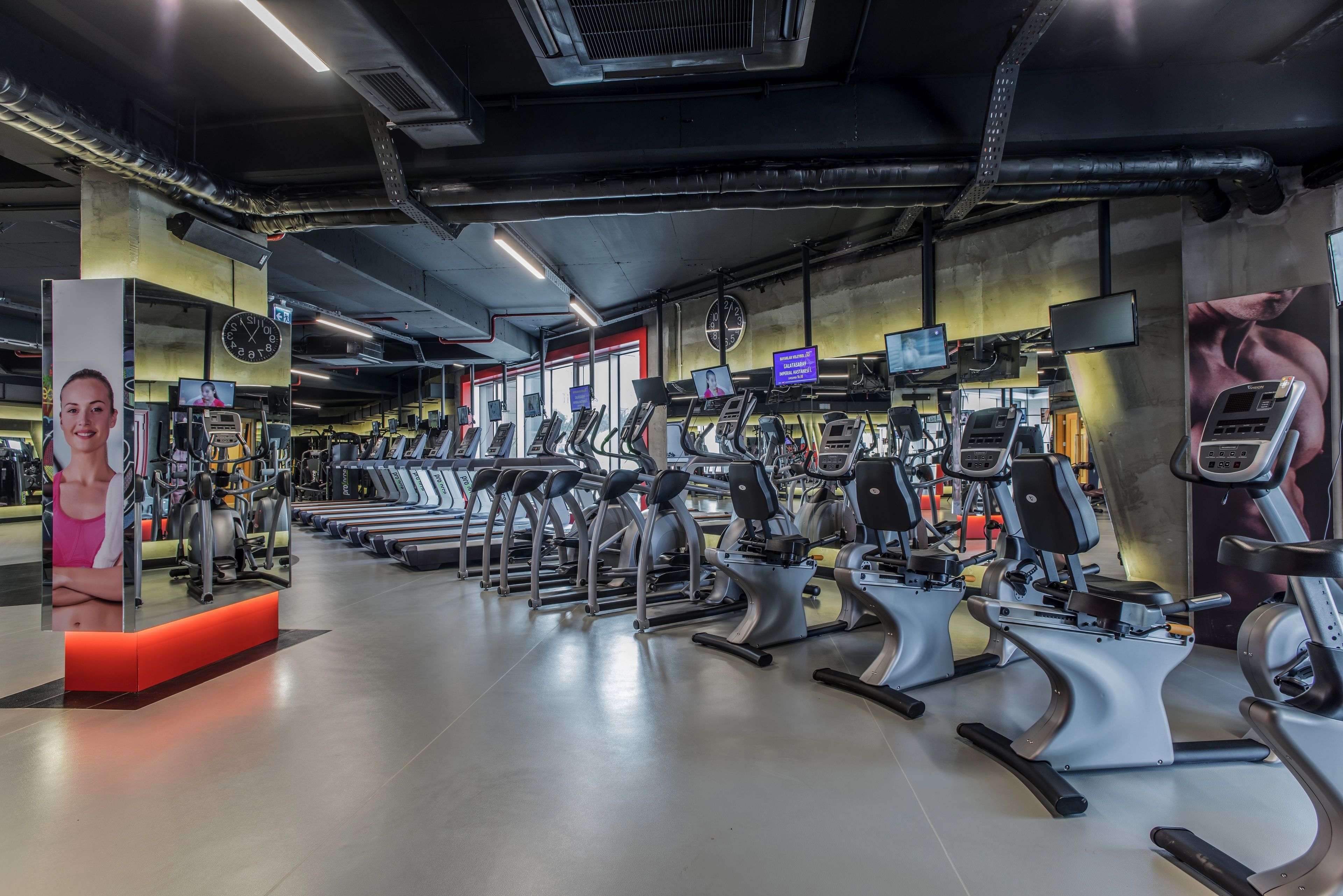Ein Fitnessstudio mit verschiedenen Cardiogeräten, einer großen Uhr und motivierenden Postern.