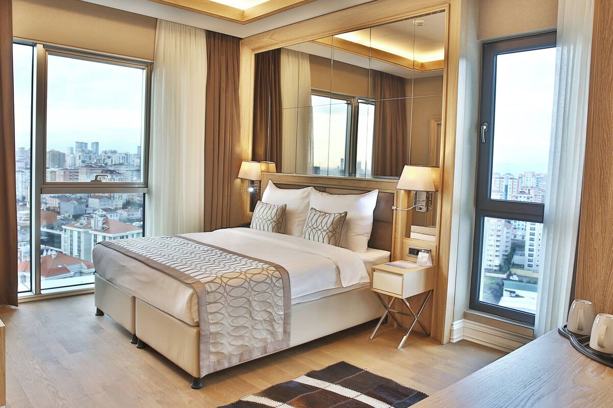 A modern hotel room with a large bed, two bedside tables, a nightstand, and a view of the city through large windows.