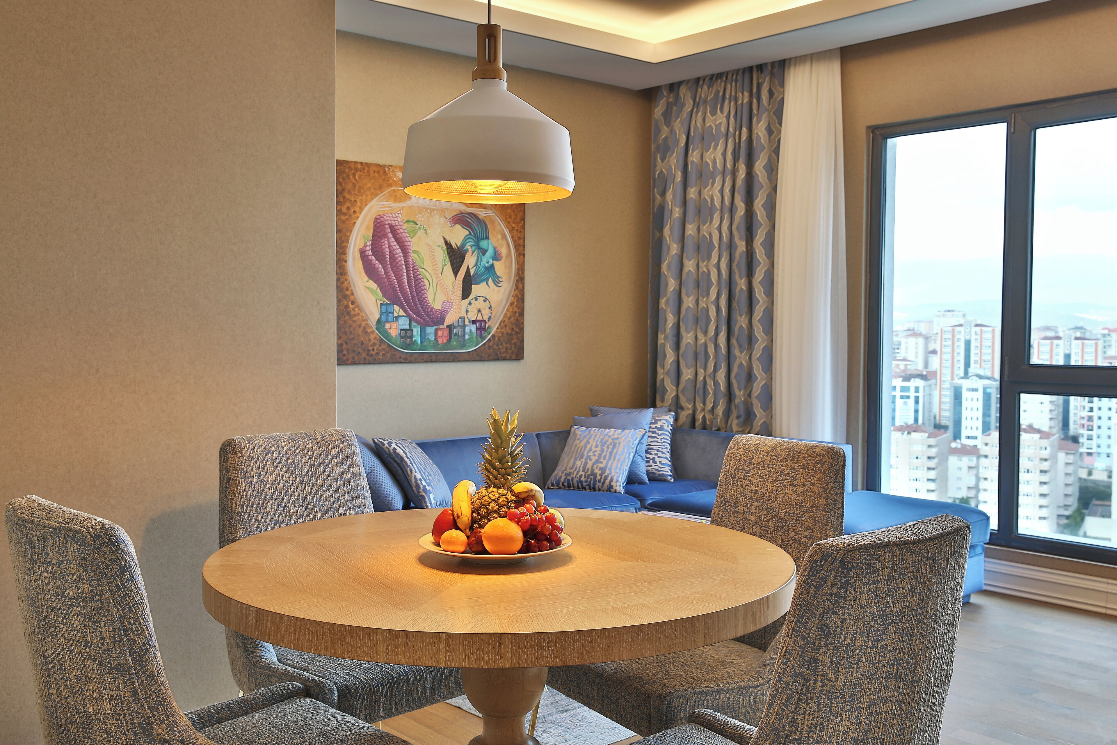 A dining area with a round table, blue upholstered chairs, a fruit bowl, and a city view through large windows.