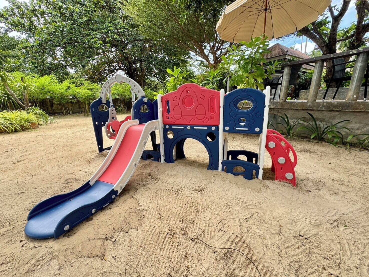 Une aire de jeux pour enfants avec des toboggans et des éléments d’escalade, installée sur un sol sablonneux, avec des arbres et un bâtiment en arrière-plan.