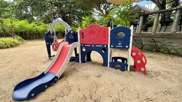 Une aire de jeux pour enfants avec des toboggans et des éléments d’escalade, installée sur un sol sablonneux, avec des arbres et un bâtiment en arrière-plan.