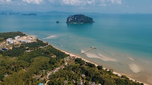 An aerial view of a coastal area with a prominent island, a pier, and a developed beachfront with buildings and greenery. - Baan Taranya Koh Yao Yai Resort (Ko Yao)