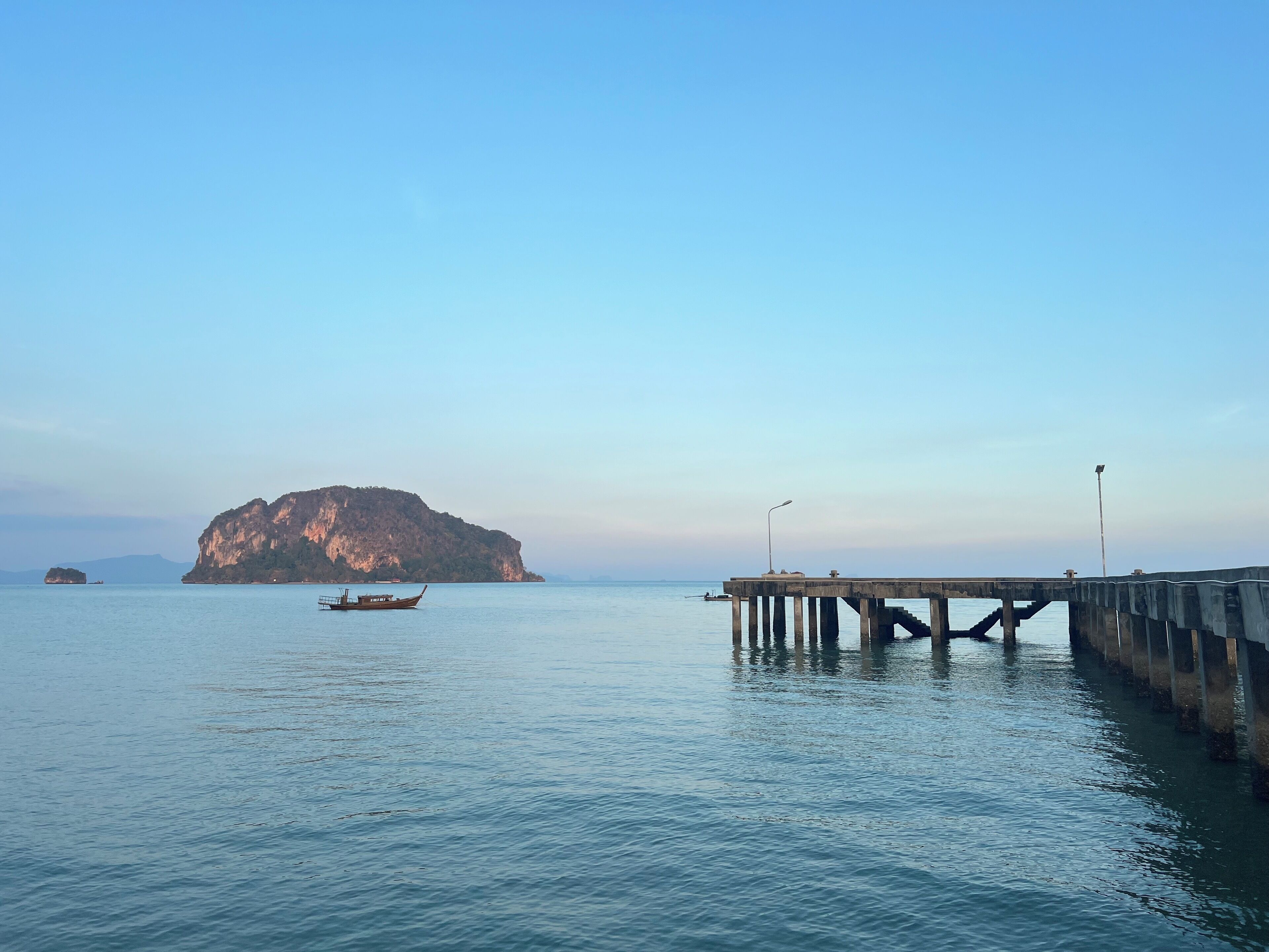 A calm sea with a pier extending into it, a boat, and a large rocky island in the background.