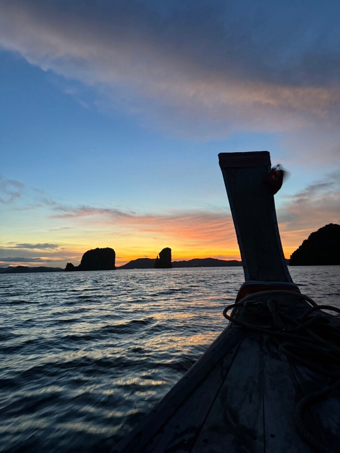 La proue d’un bateau, à laquelle est attachée une corde, est orientée vers un coucher de soleil sur une étendue d’eau calme, avec des formations rocheuses visibles au loin.