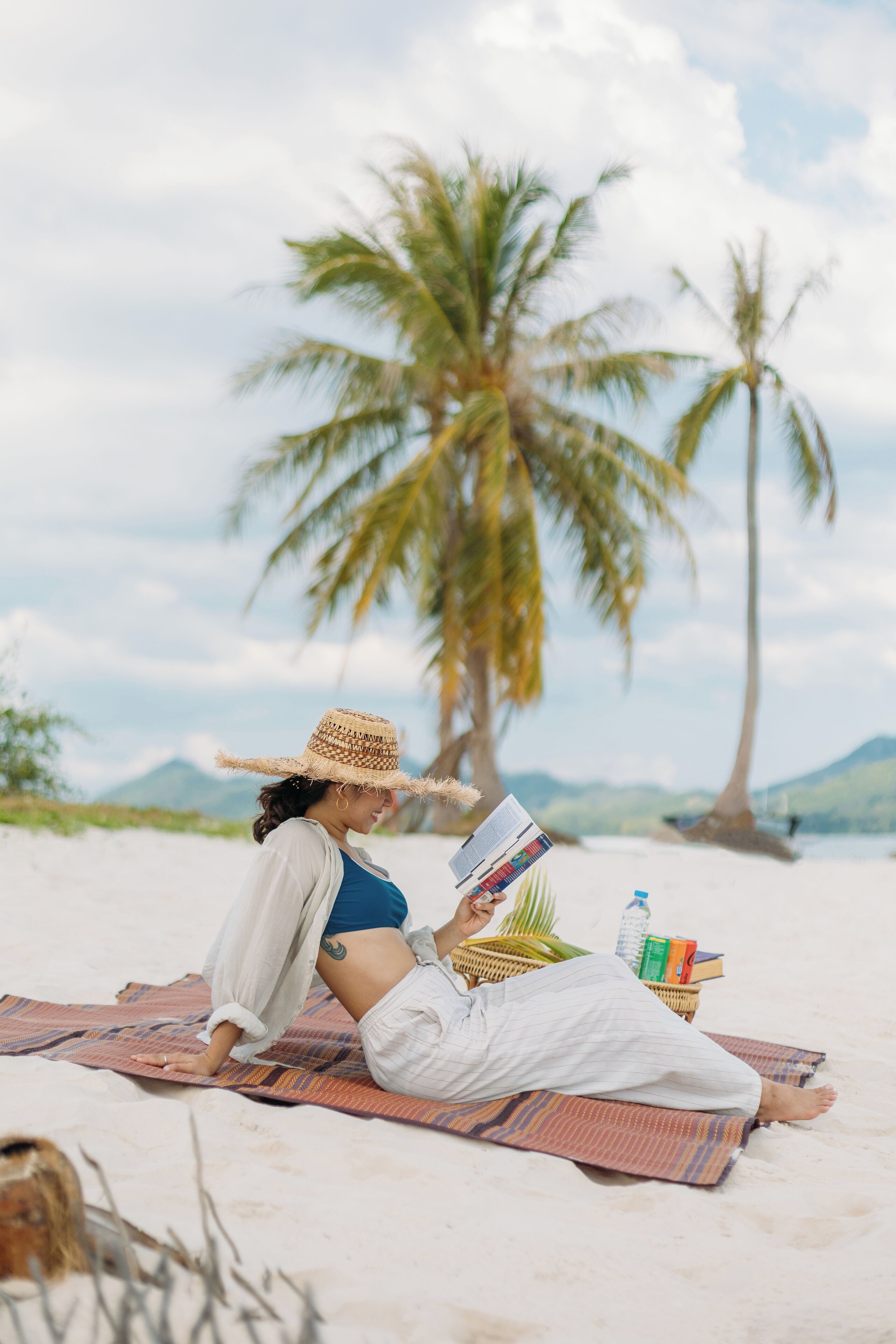 Una persona recostada sobre una toalla en la playa, leyendo un libro, con una palmera y montañas al fondo.