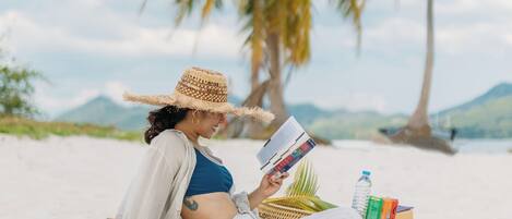 Una persona recostada sobre una toalla en la playa, leyendo un libro, con una palmera y montañas al fondo.