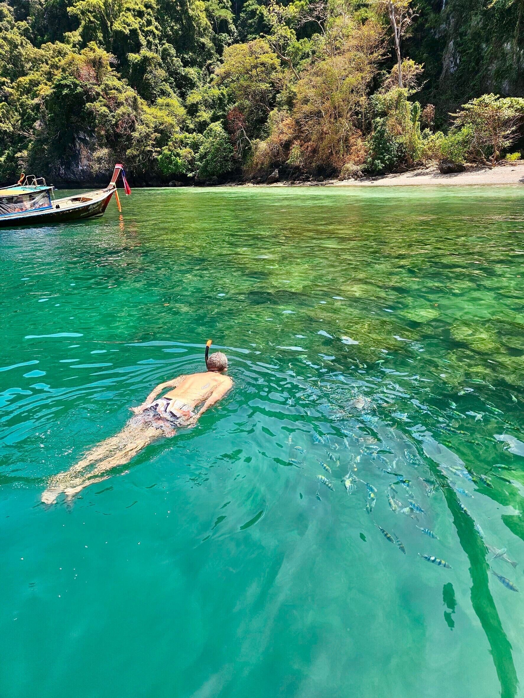 A person swimming in clear turquoise water with a boat and forested shore in the background.