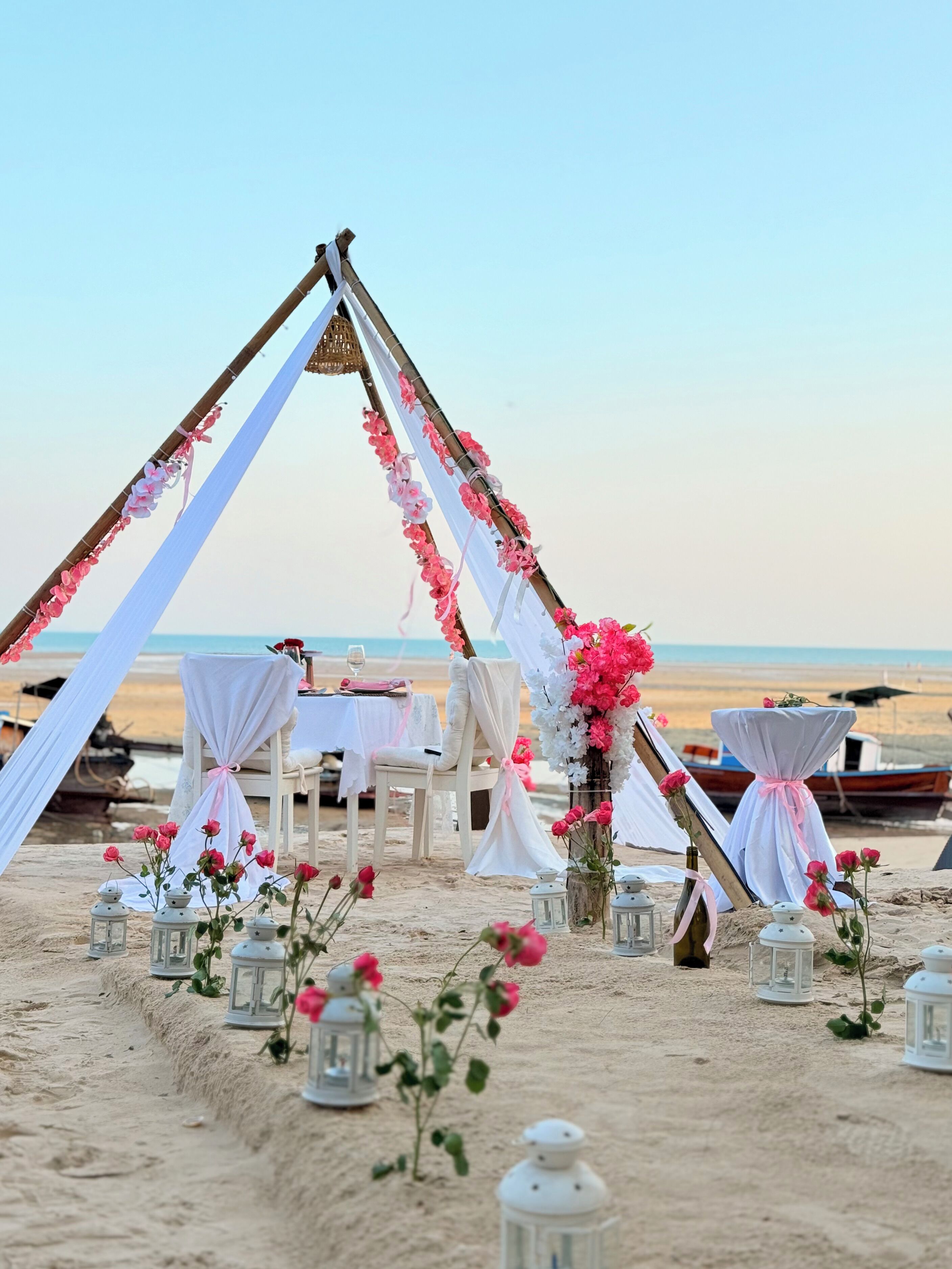 Une cérémonie de mariage organisée sur la plage, avec une arche en bois, des draperies blanches et des fleurs roses. Les tables sont dressées avec des nappes blanches et des chaises.