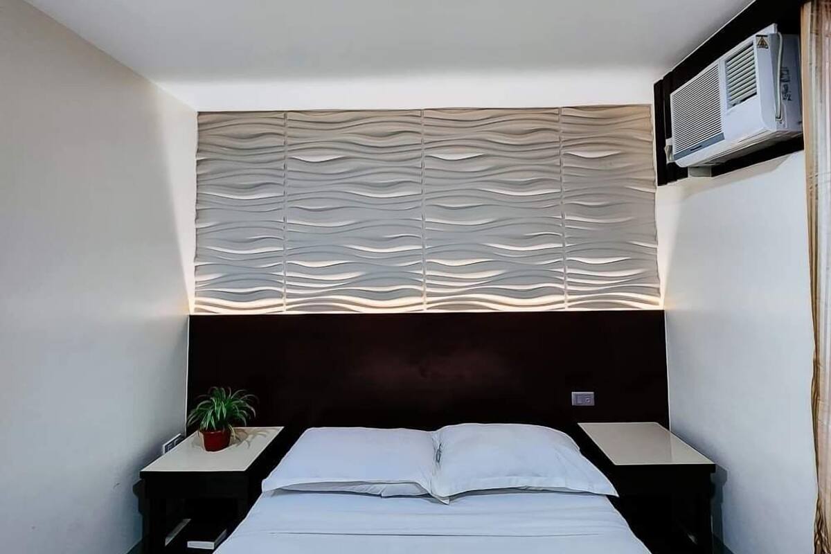 A bedroom with a bed, a nightstand, a potted plant, and a wall-mounted air conditioner.