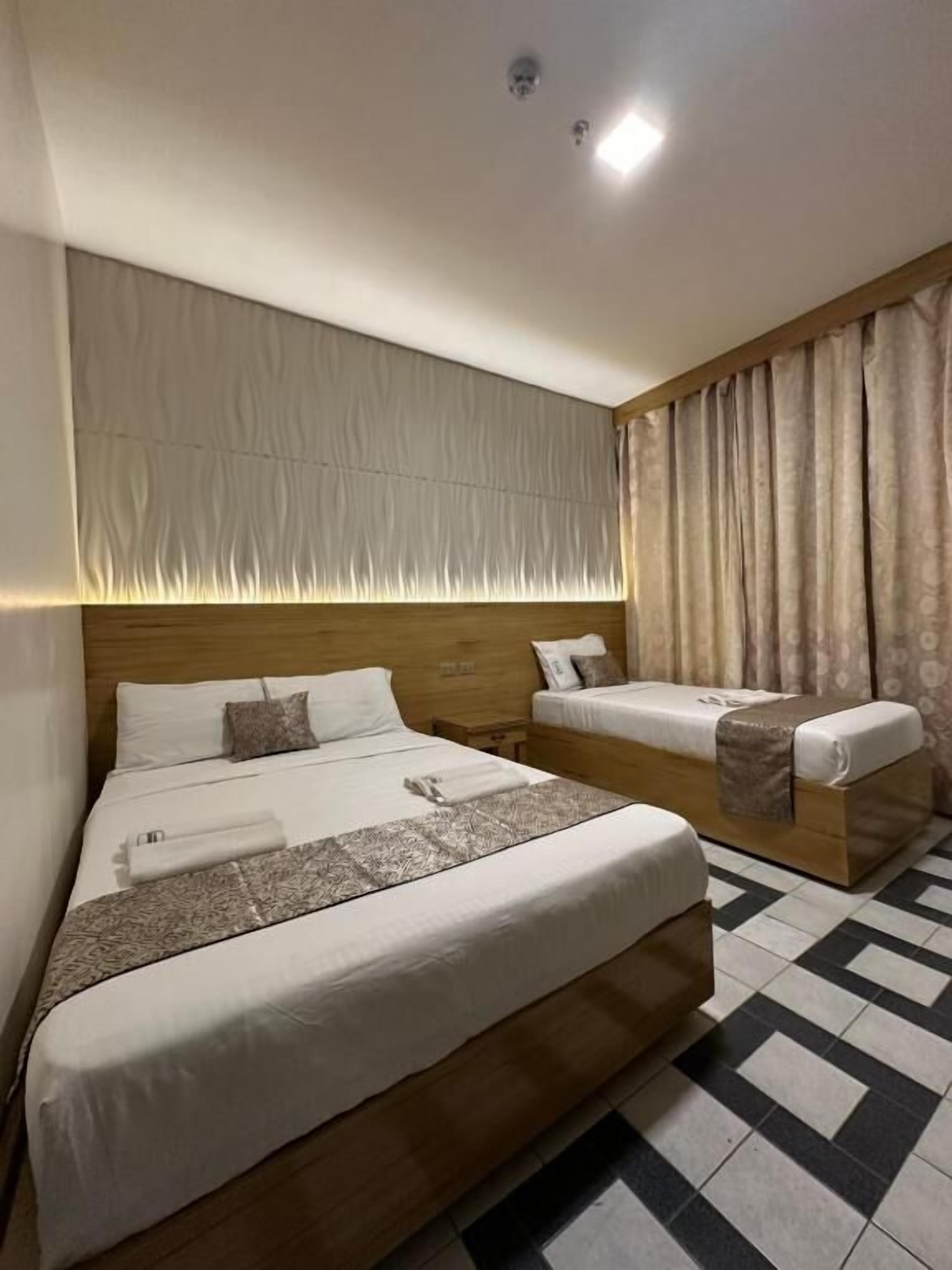 Una habitación de hotel con una cama grande, otra más pequeña, un cabecero, una mesita de noche y una cortina.