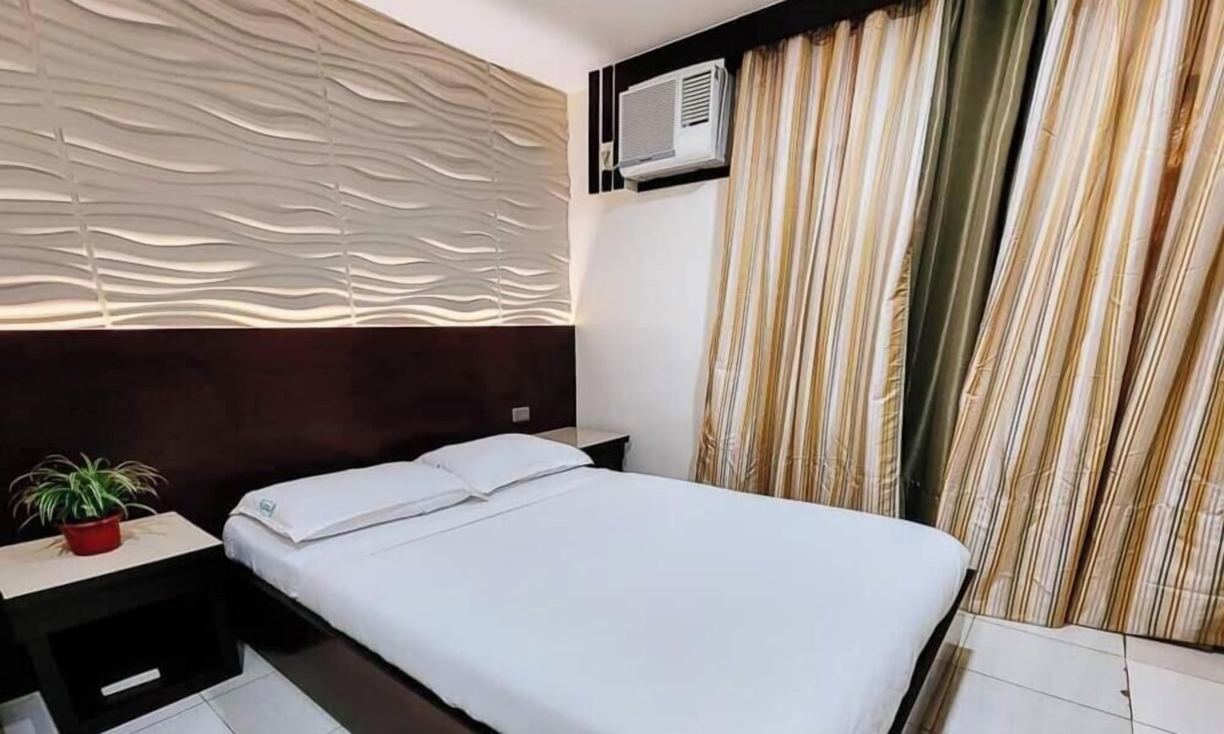 A hotel room with a bed, a nightstand, a potted plant, a wall-mounted air conditioner, and a window with curtains.