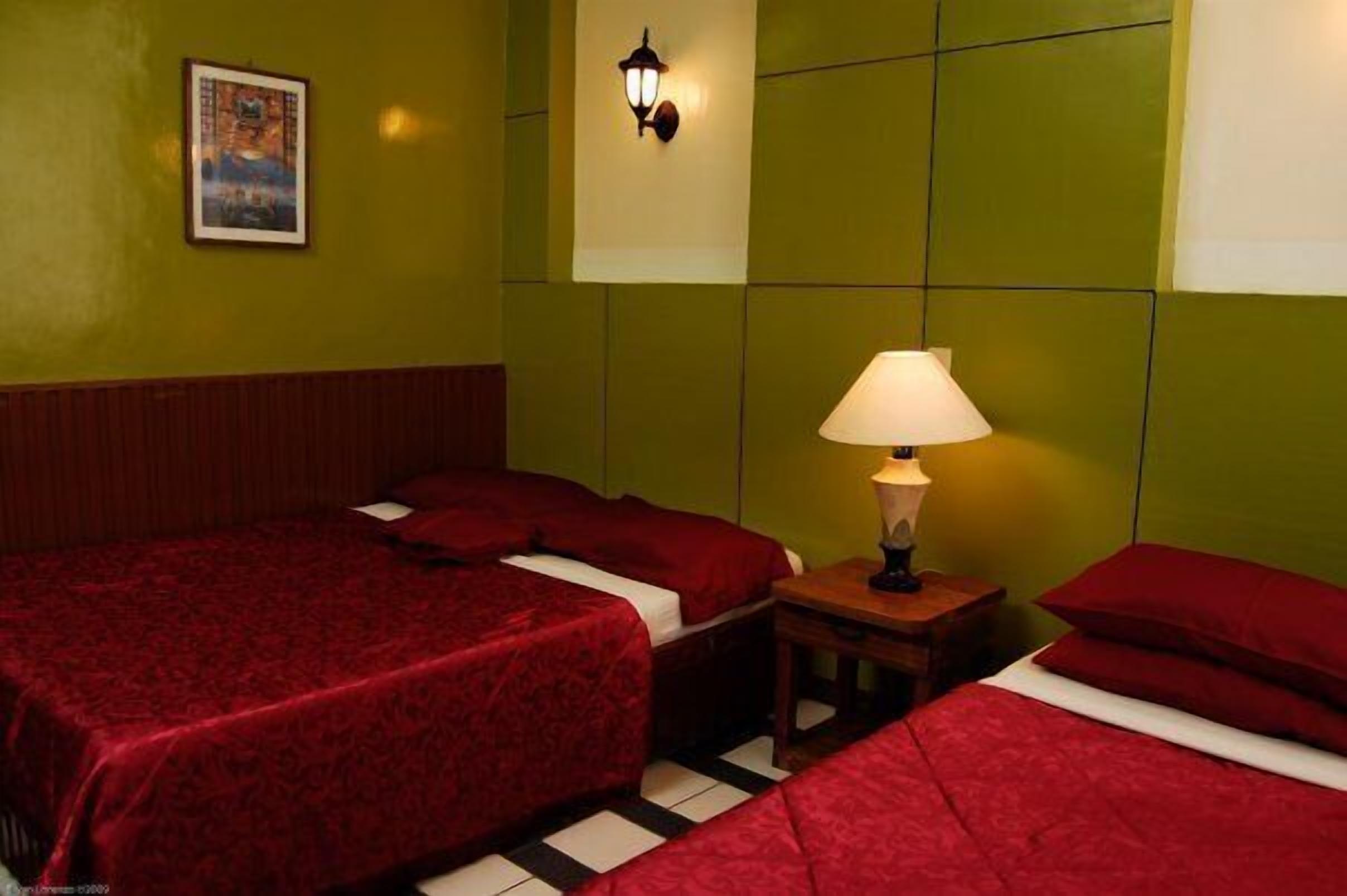 Habitación de hotel con dos camas, una mesita de noche con lámpara, una lámpara de pared e imagen enmarcada.