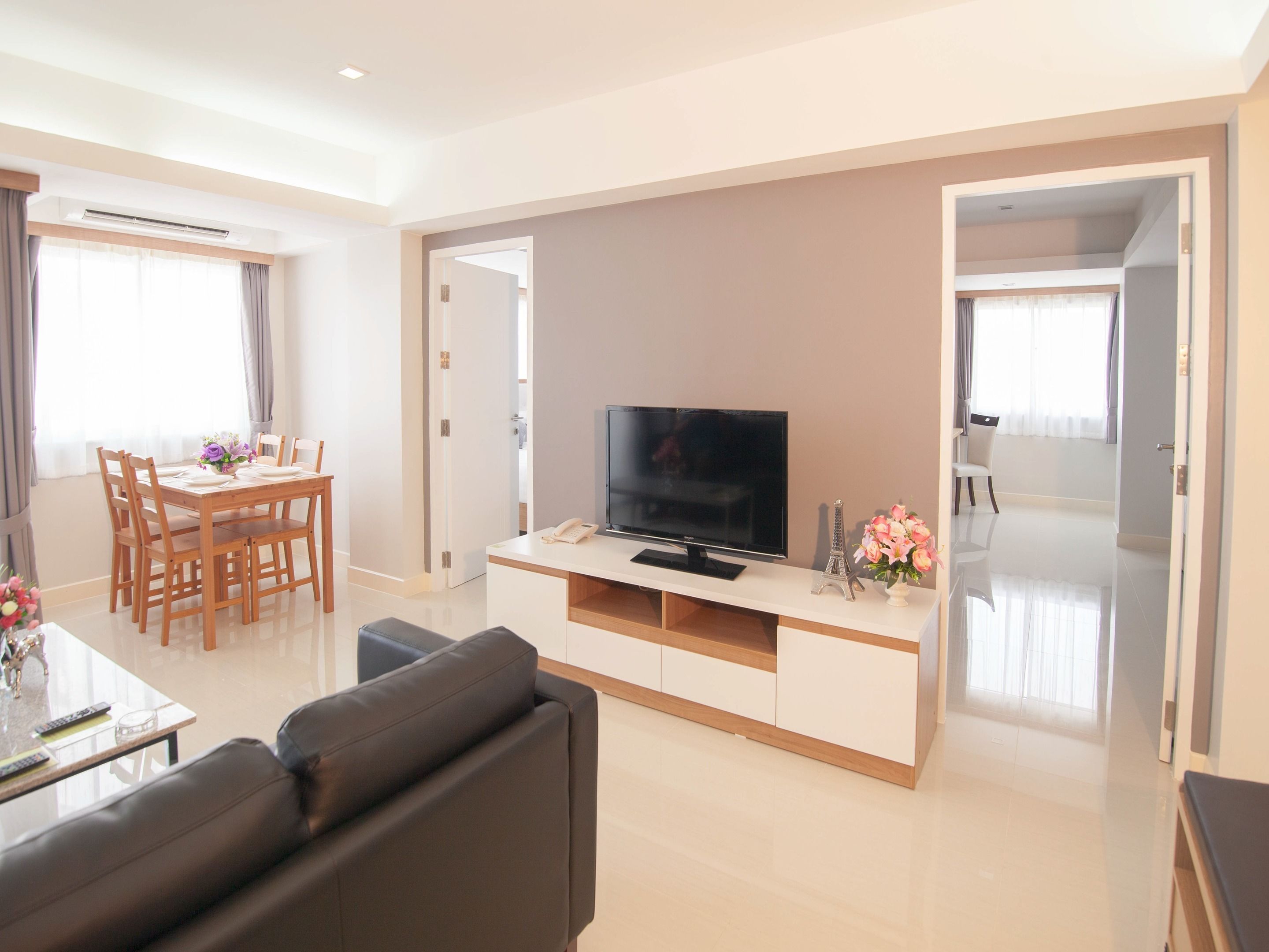 A modern living room with a flat-screen TV, a sofa, a dining table with chairs, and a small table with a vase of flowers.