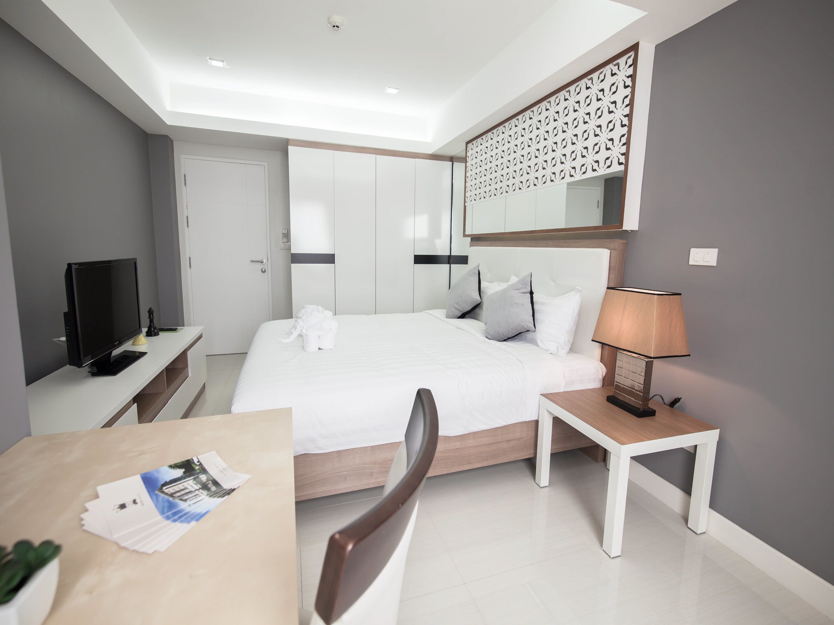 Ein modernes Hotelzimmer mit Bett, Nachttisch, Lampe, Fernseher und Schreibtisch mit Stuhl und einigen Papieren.