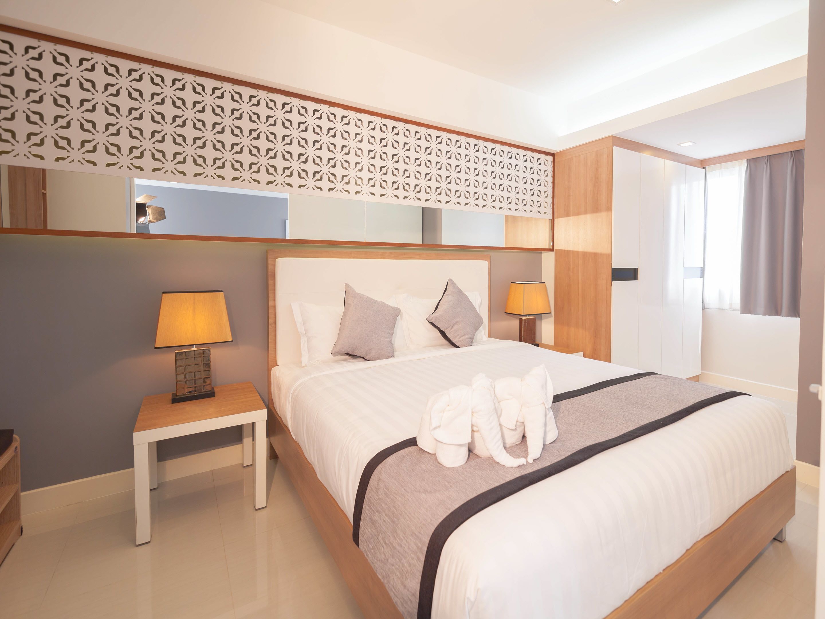 Habitación de hotel con una cama bien hecha, dos lámparas de noche, un cabecero de madera, un panel decorativo en la pared y un gran ventanal con cortinas.