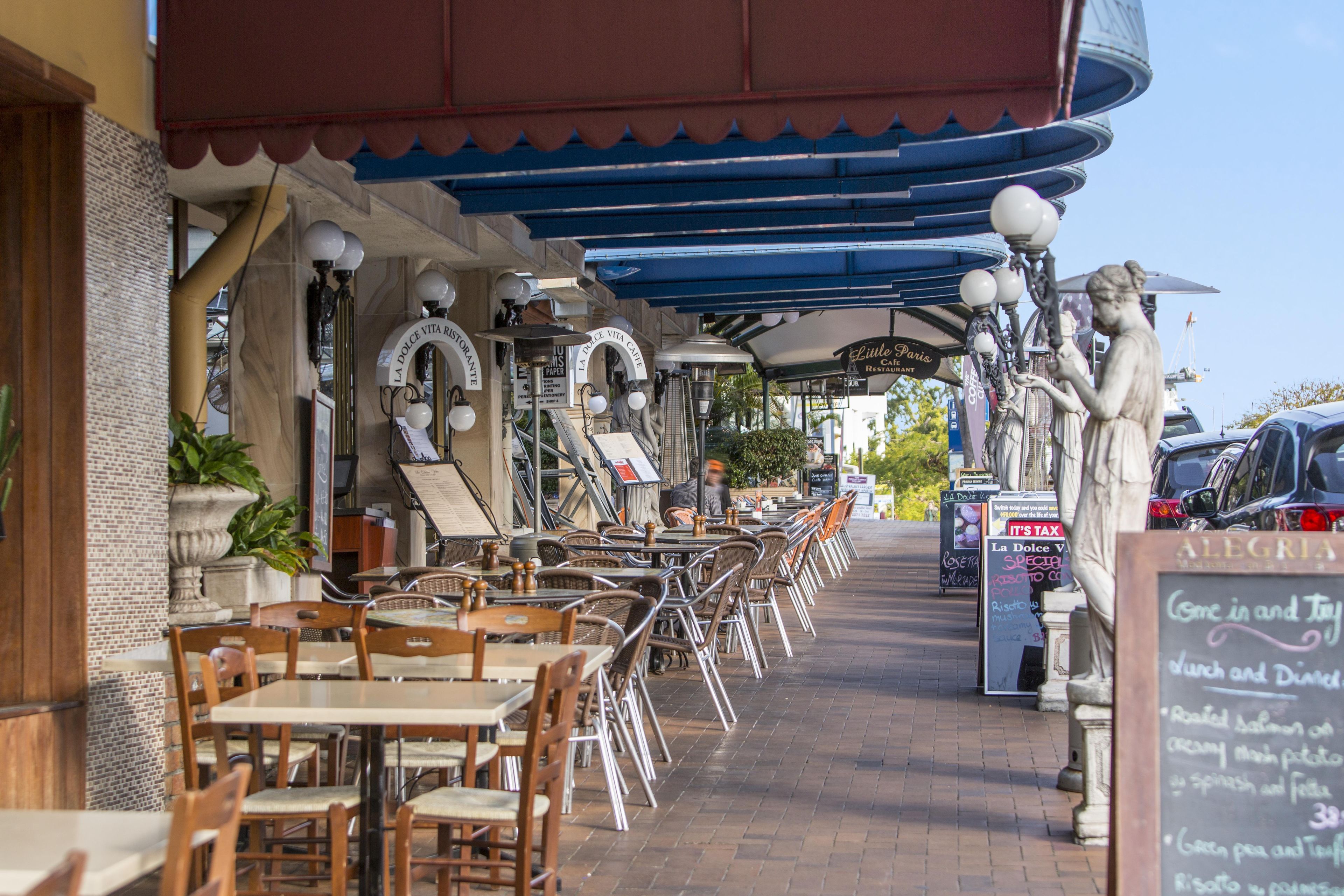 Ett utomhuskafé med bord och stolar, ett takförsett sittområde och en skylt med texten ”Lilla Paris-restaurangen”.