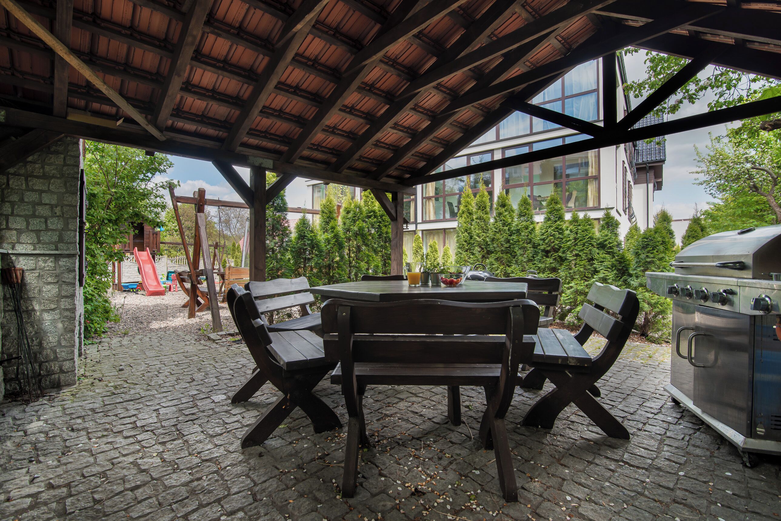 An outdoor patio with a wooden table and benches, a grill, and a playground in the background.