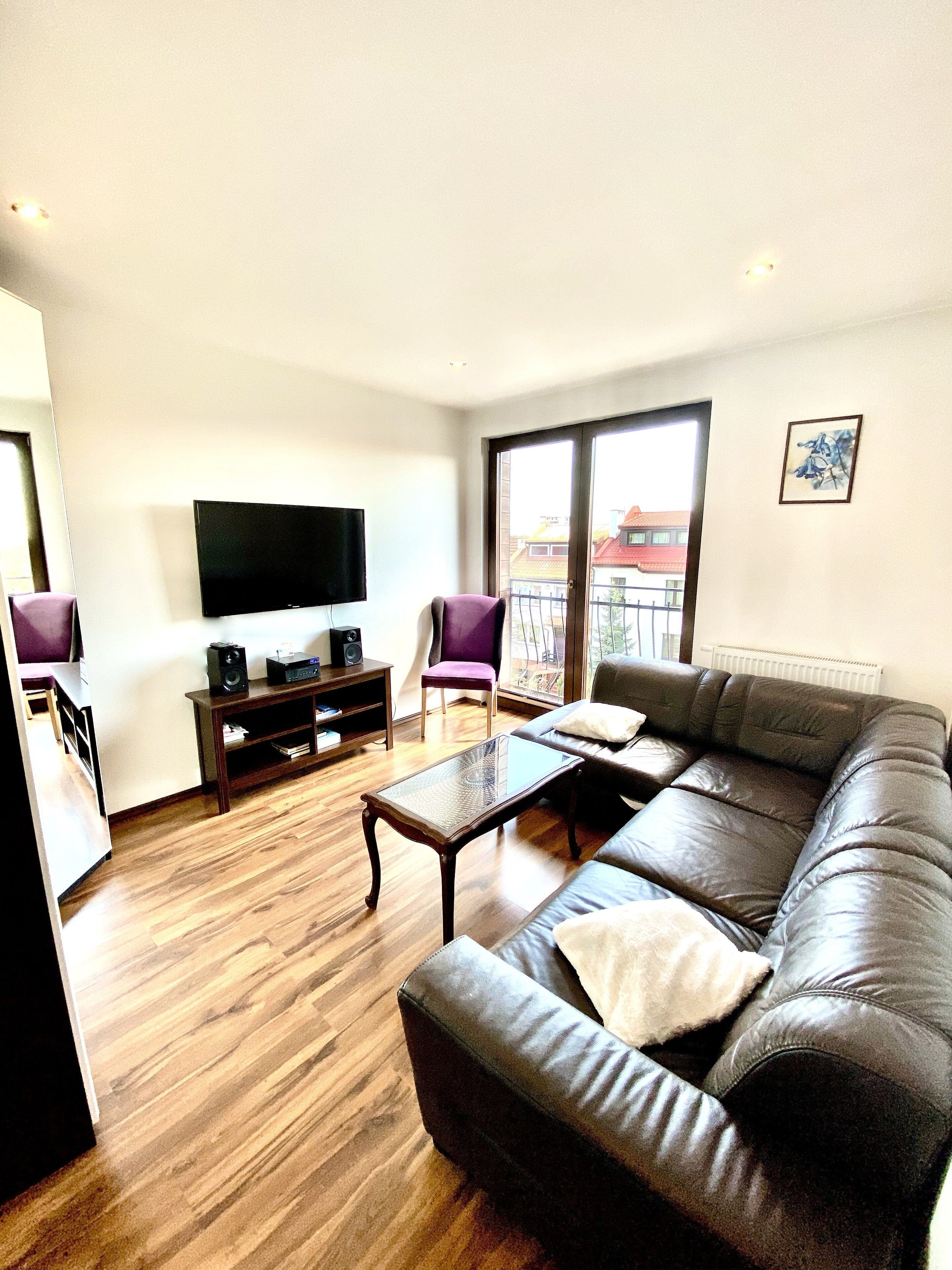 A modern living room with a leather sofa, a glass coffee table, a flat-screen TV, and a balcony with a view.