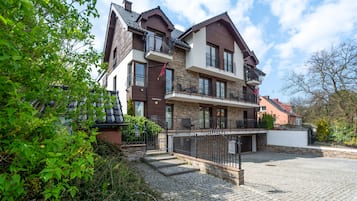 Traditional two-story building with a brick and stone facade, featuring a front entrance and balconies on the upper floor, set amidst lush greenery.