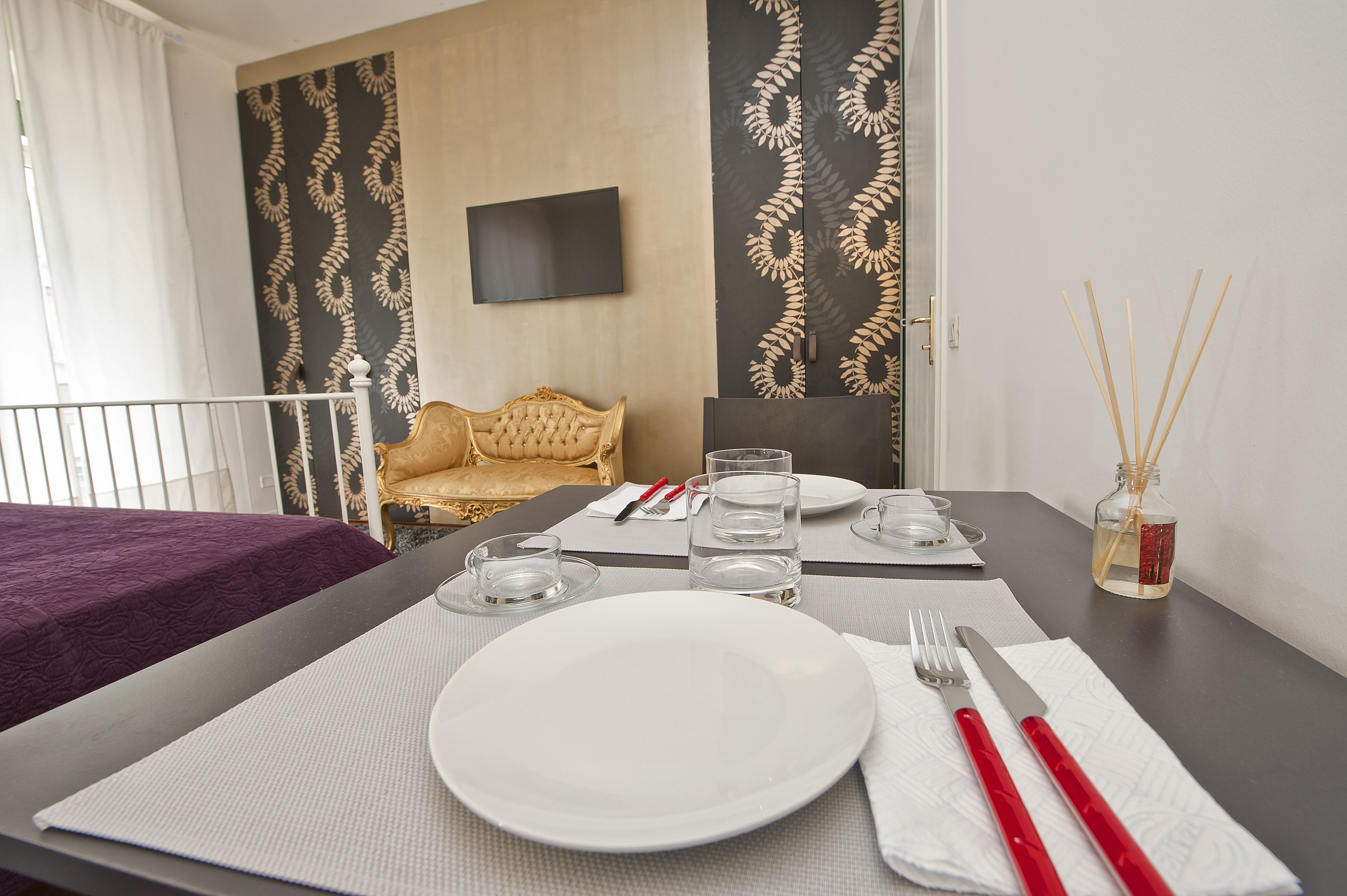A neatly set dining table with a white plate, cutlery, and glasses, a small bottle of water, and a decorative reed diffuser.