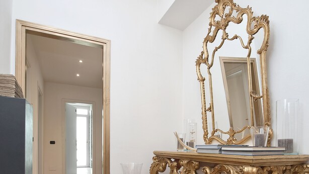 A room with a wooden floor, a gold ornate table with a mirror, and a door leading to another room. - Verona Maison (Verona)
