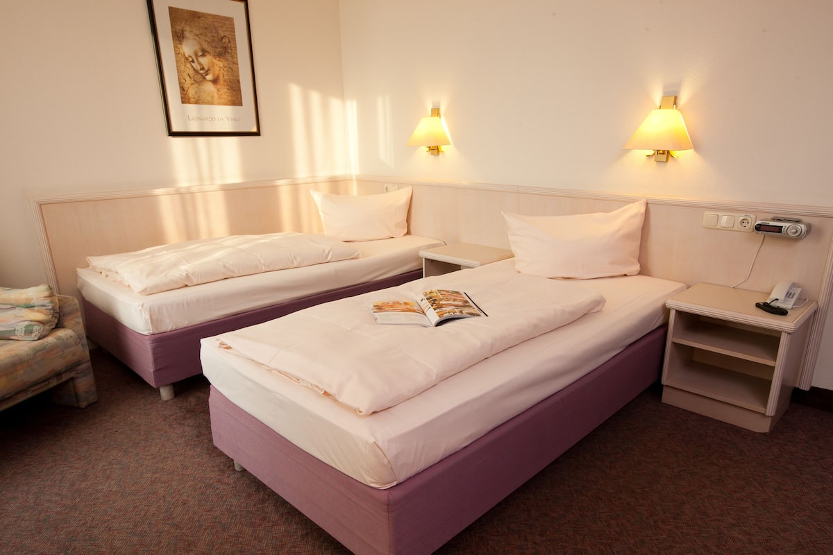 A hotel room with two beds, a bedside table with a lamp, a chair, and a painting on the wall.