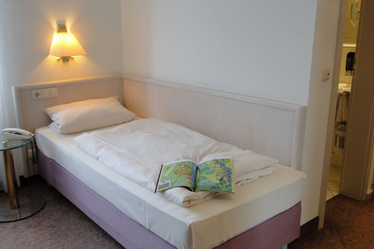 A single bed with a white headboard and a bedside table with a lamp. A book is placed on the bed.