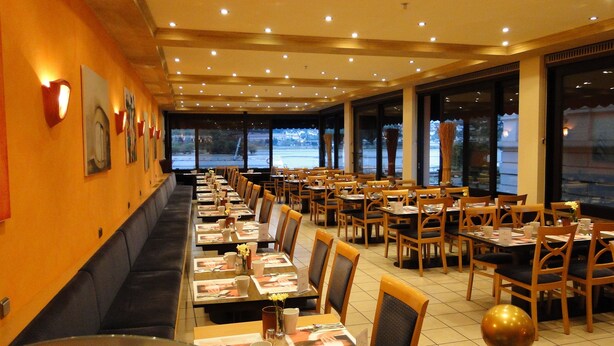 A restaurant with long tables, wooden chairs, and a view of the outside through large windows. - Hoteltraube Rudesheim (Ruedesheim am Rhein)