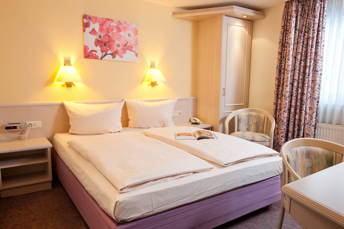 A hotel room with two beds, a desk, a chair, a wardrobe, and a painting on the wall.