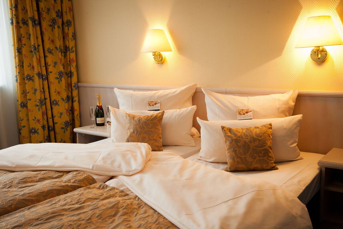 A hotel room with a bed, pillows, a nightstand, a bottle of champagne, and a floral curtain.