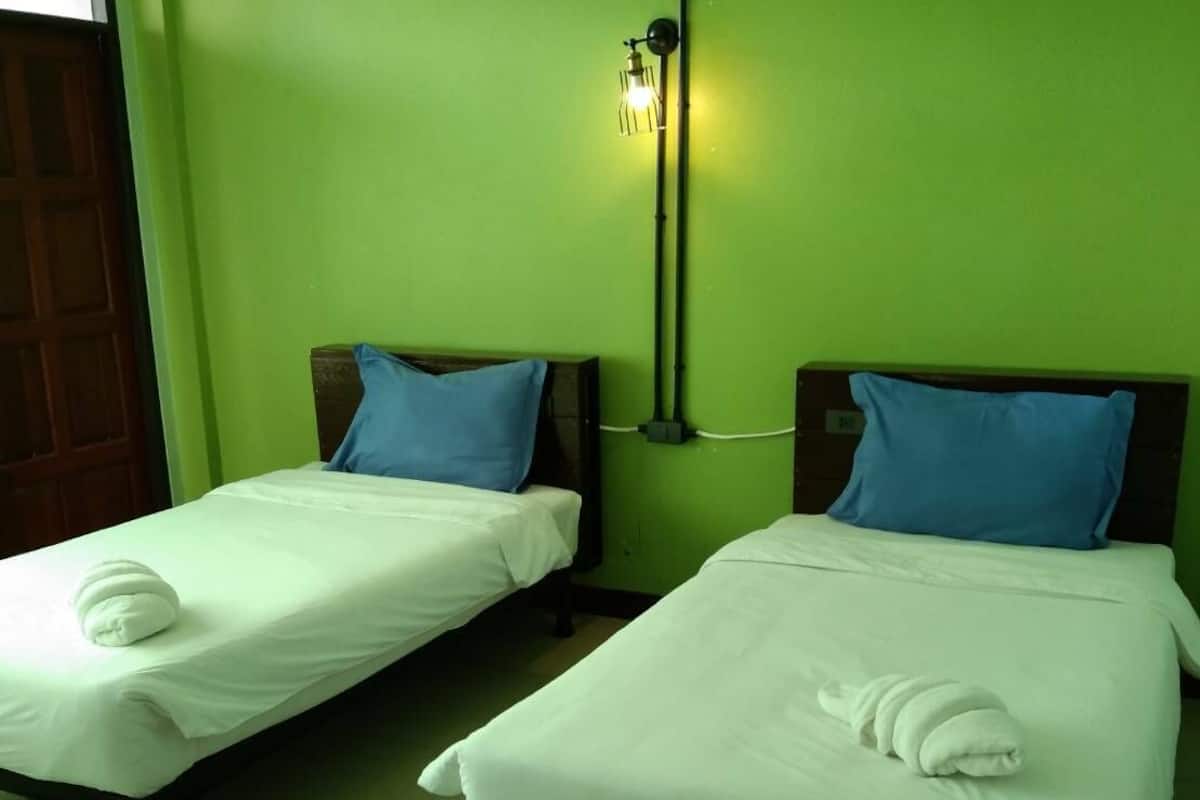 A hotel room with two single beds, each with a white mattress and blue pillows, and a green wall.