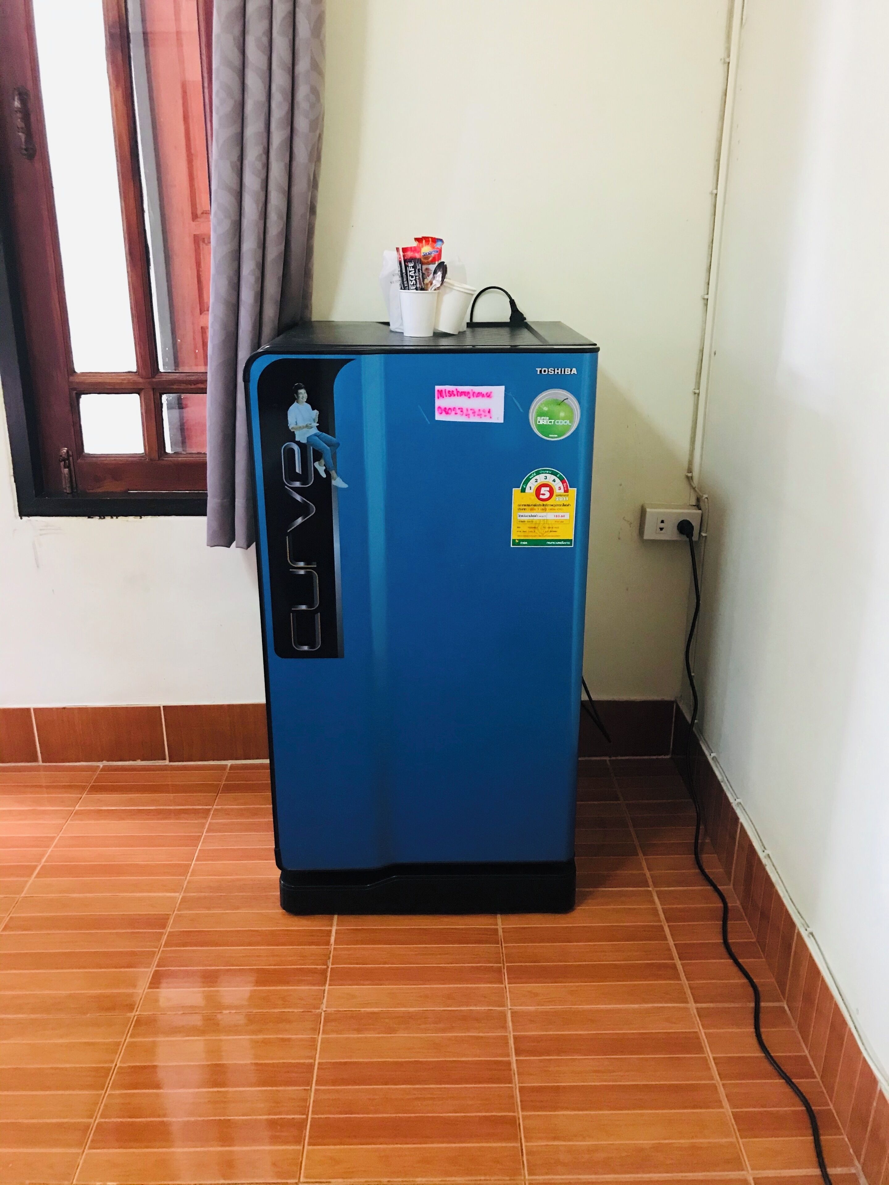 Un refrigerador azul con calcomanías, una ventana con cortinas y un piso de baldosas.