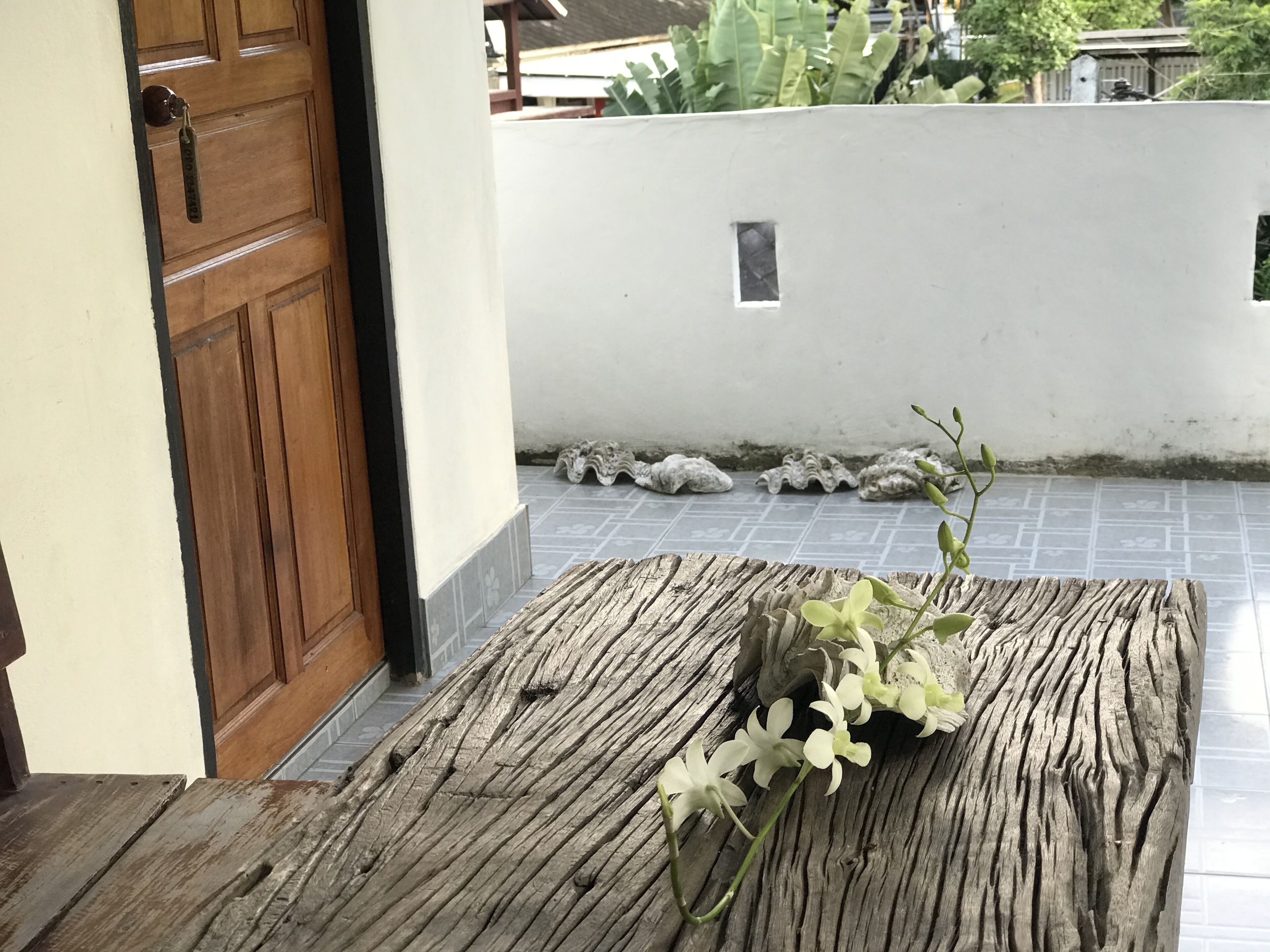Una mesa de madera con un jarrón de flores blancas en un umbral que da a un patio con pared blanca y suelo de baldosas.