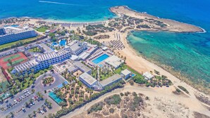 An aerial view of a resort with a large pool, multiple buildings, and a beach area. - Dome Beach Marina Hotel & Resort (Ayia Napa)