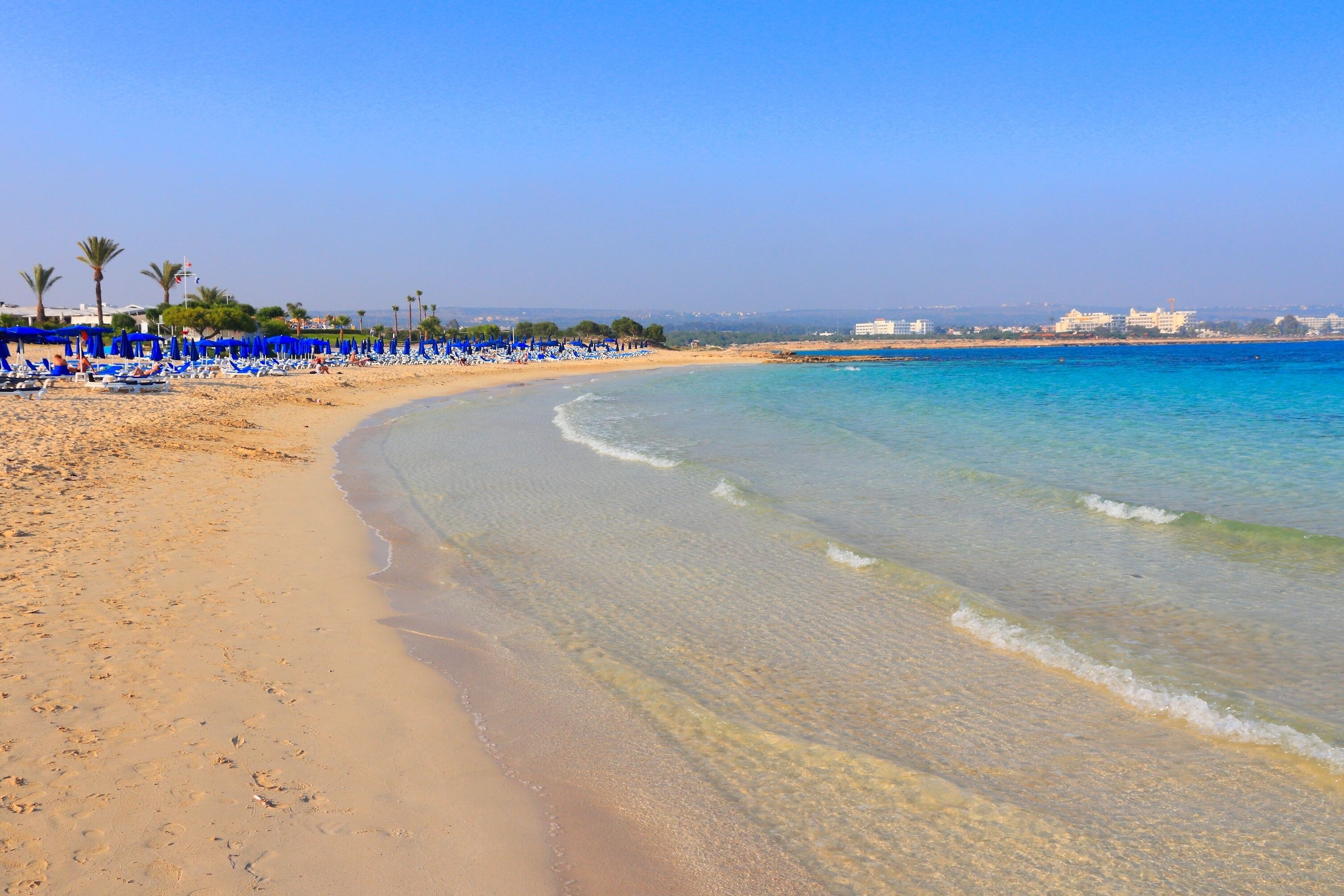Una playa con aguas azul cristalinas, orilla arenosa y sombrillas azules.