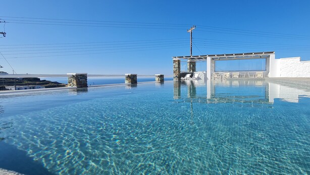 Una piscina infinita cristalina con pilares de piedra, una pérgola y sillas de descanso, con vistas a la costa.