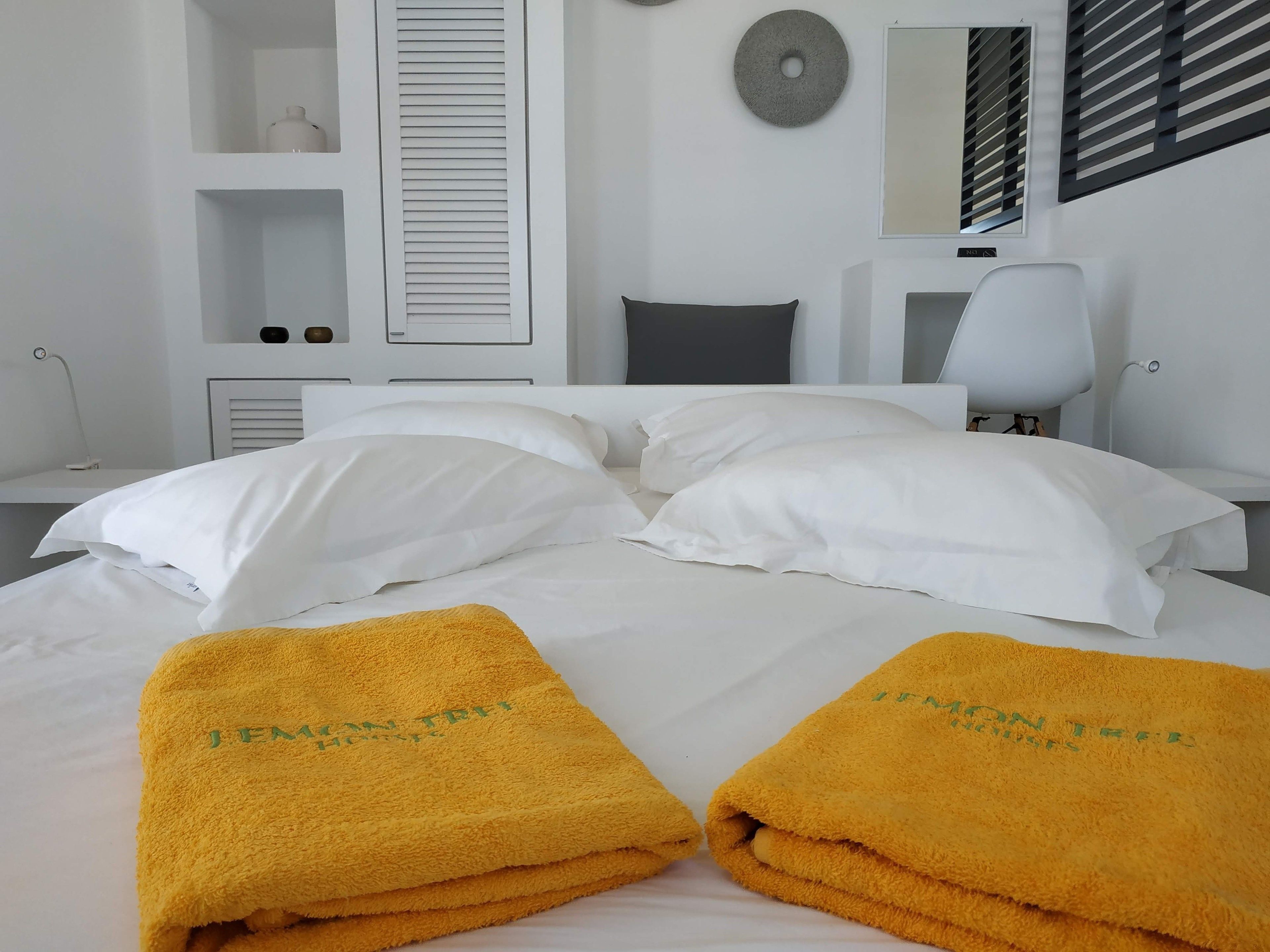 Una habitació d'hotel amb un llit, almoines blanques, una taula de nit, una làmpada i una tovallola amb el text "JEMAN TREE" brodat.