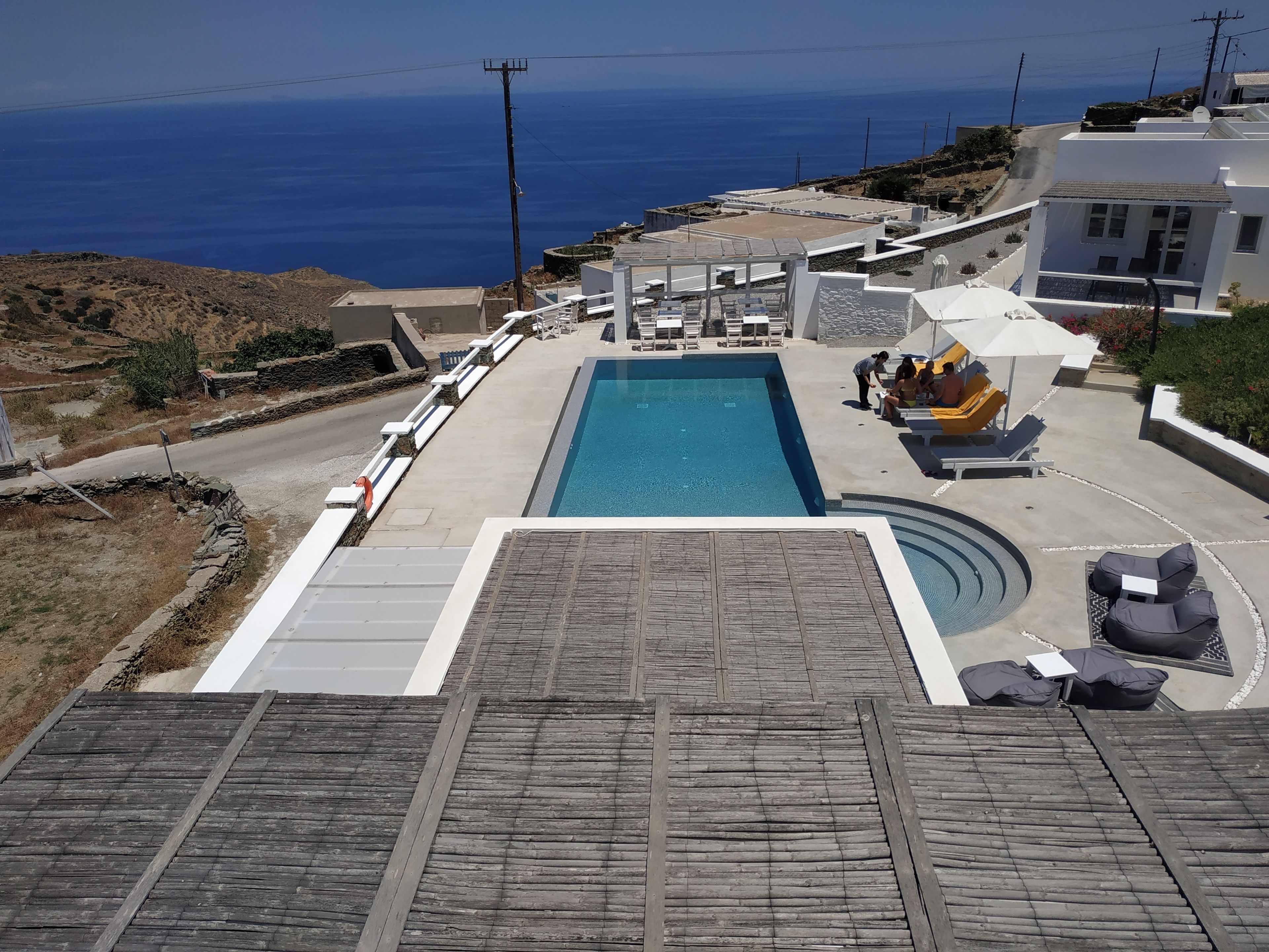 Zona de piscina con vista al mar, mobiliario exterior y una carretera que lleva a un edificio blanco.