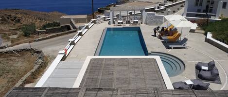 Zona de piscina con vista al mar, mobiliario exterior y una carretera que lleva a un edificio blanco.