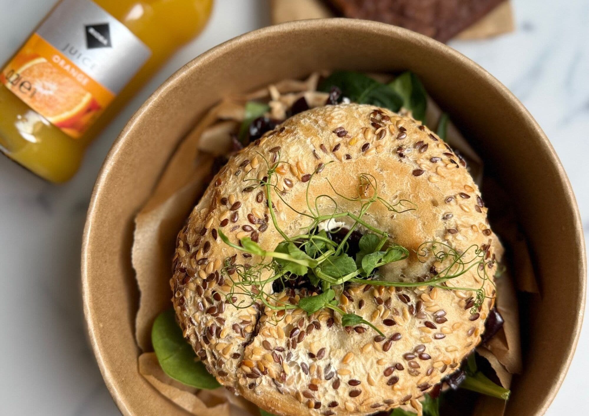 Een sandwich in een kartonnen verpakking, belegd met zaden, samen met een fles sinaasappelsap.