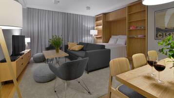 A modern hotel room with a sofa, a dining table with chairs, a TV, and a bookshelf.
