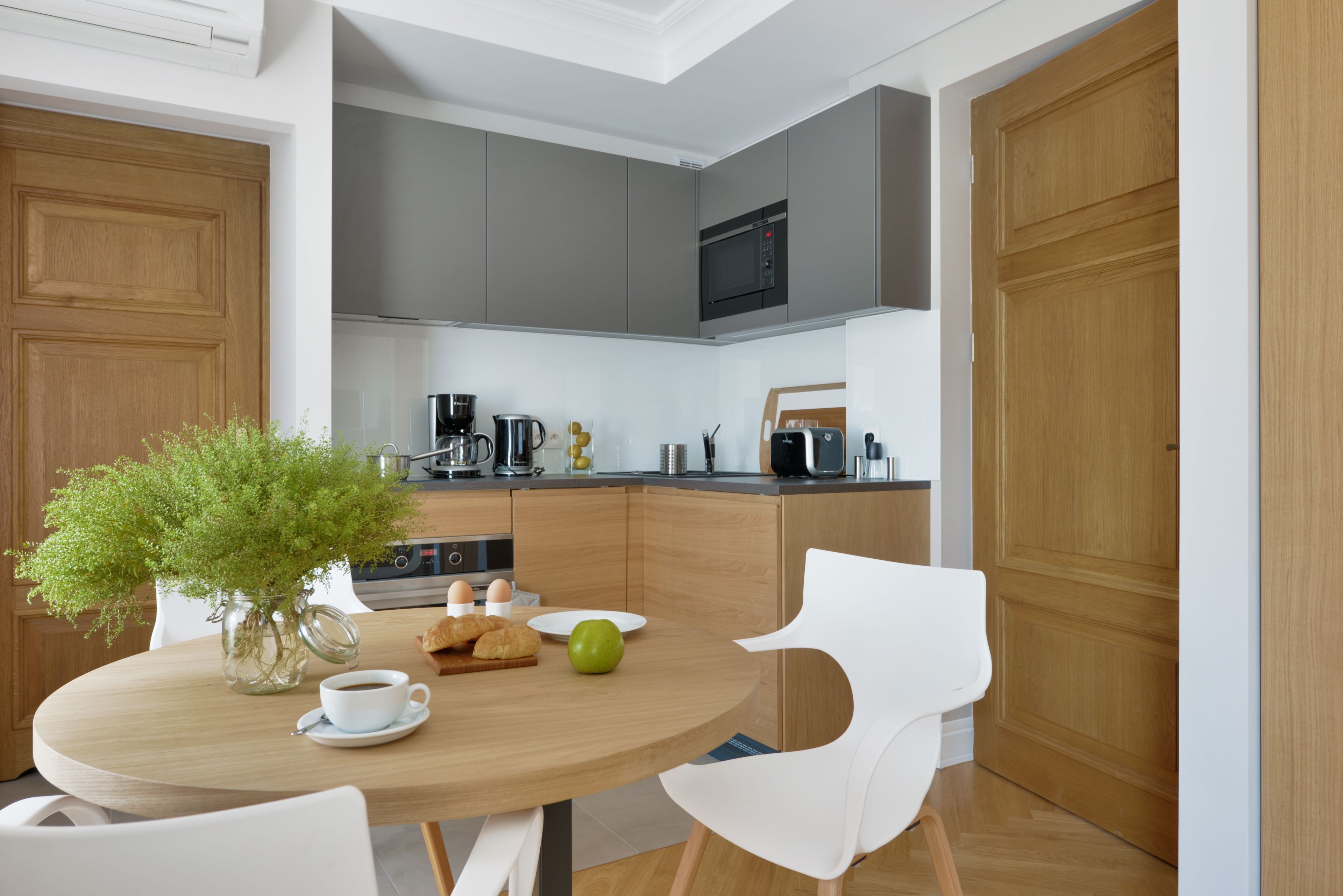 A modern kitchen with grey cabinets, a microwave, and a coffee maker. There is a dining table with chairs and a vase of green plants.