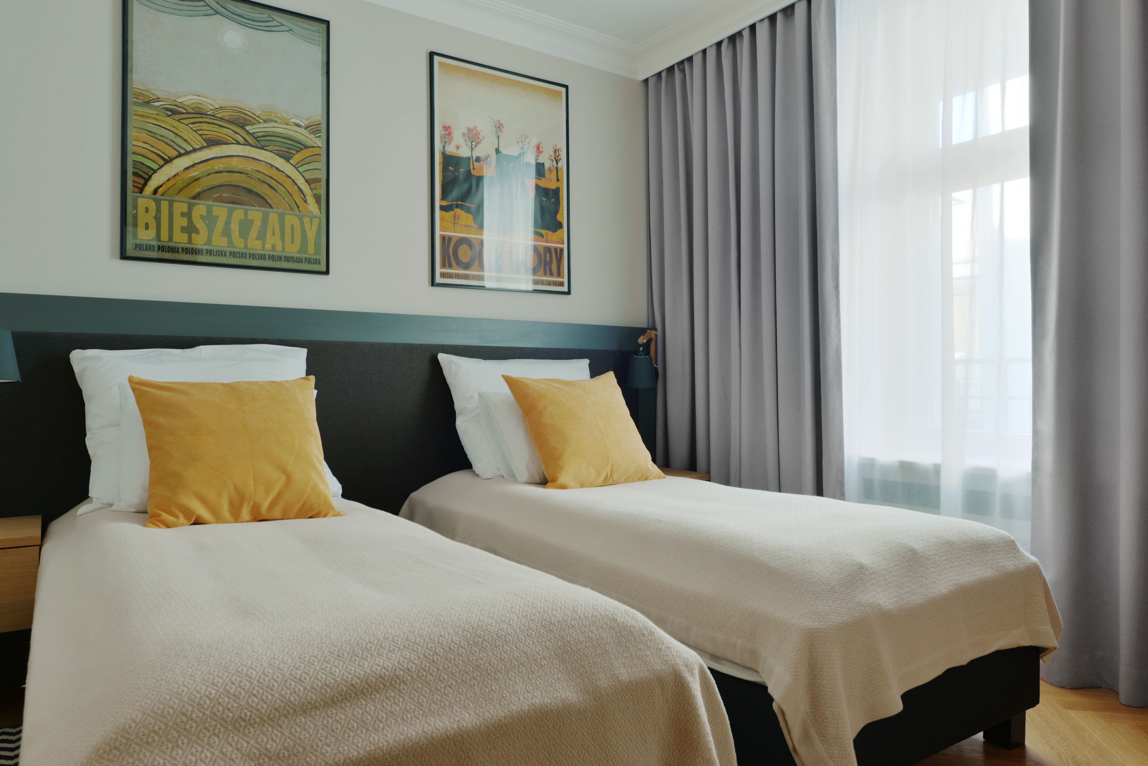 Une chambre d’hôtel avec deux lits, chacun agrémenté d’un oreiller blanc et jaune, d’une table de chevet en bois et d’une grande fenêtre avec des rideaux gris.