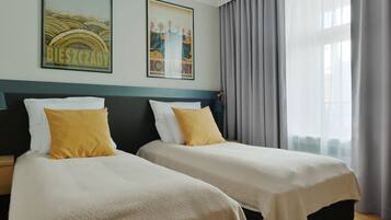 Une chambre d’hôtel avec deux lits, chacun agrémenté d’un oreiller blanc et jaune, d’une table de chevet en bois et d’une grande fenêtre avec des rideaux gris.