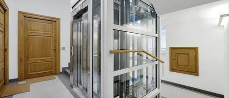 A modern interior with a glass elevator, wooden doors, and a tiled floor.