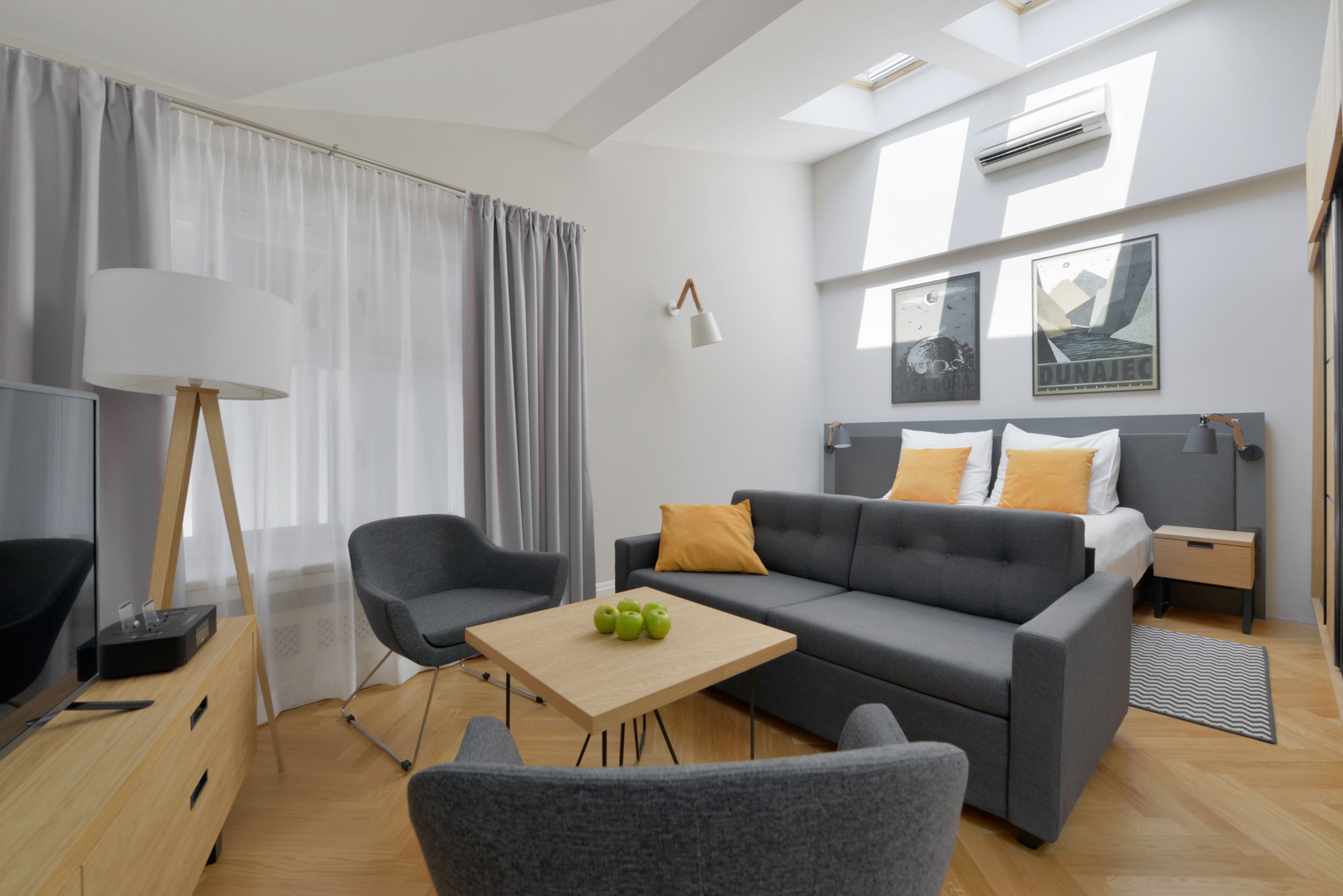 A modern living room with a grey sofa, a wooden coffee table with green apples, a grey armchair, a floor lamp, and a TV on a wooden cabinet.