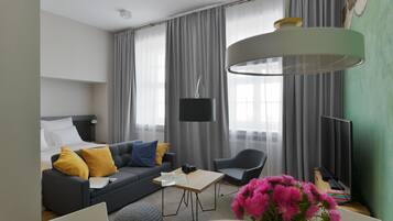 A modern hotel room with a grey sofa, a small table, a TV, and a large window with curtains.