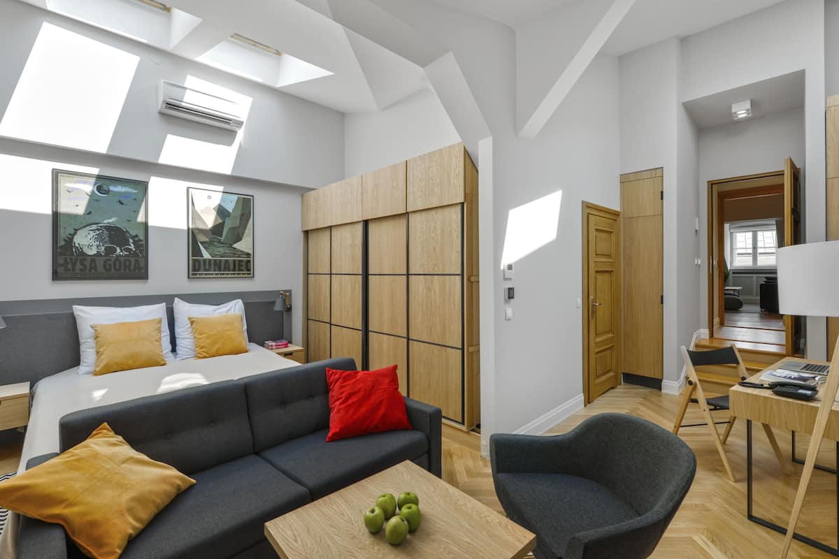Una habitación de hotel moderna con cama, sofá gris, almohada roja, armario de madera y escritorio con lámpara y manzanas.